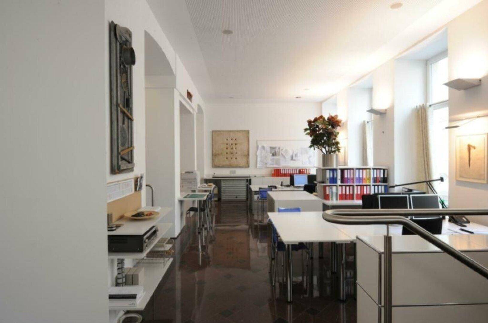 Weiterer Blick in das offene Büro mit Schreibtischen, Kunstwerken an den Wänden und hohen Decken.