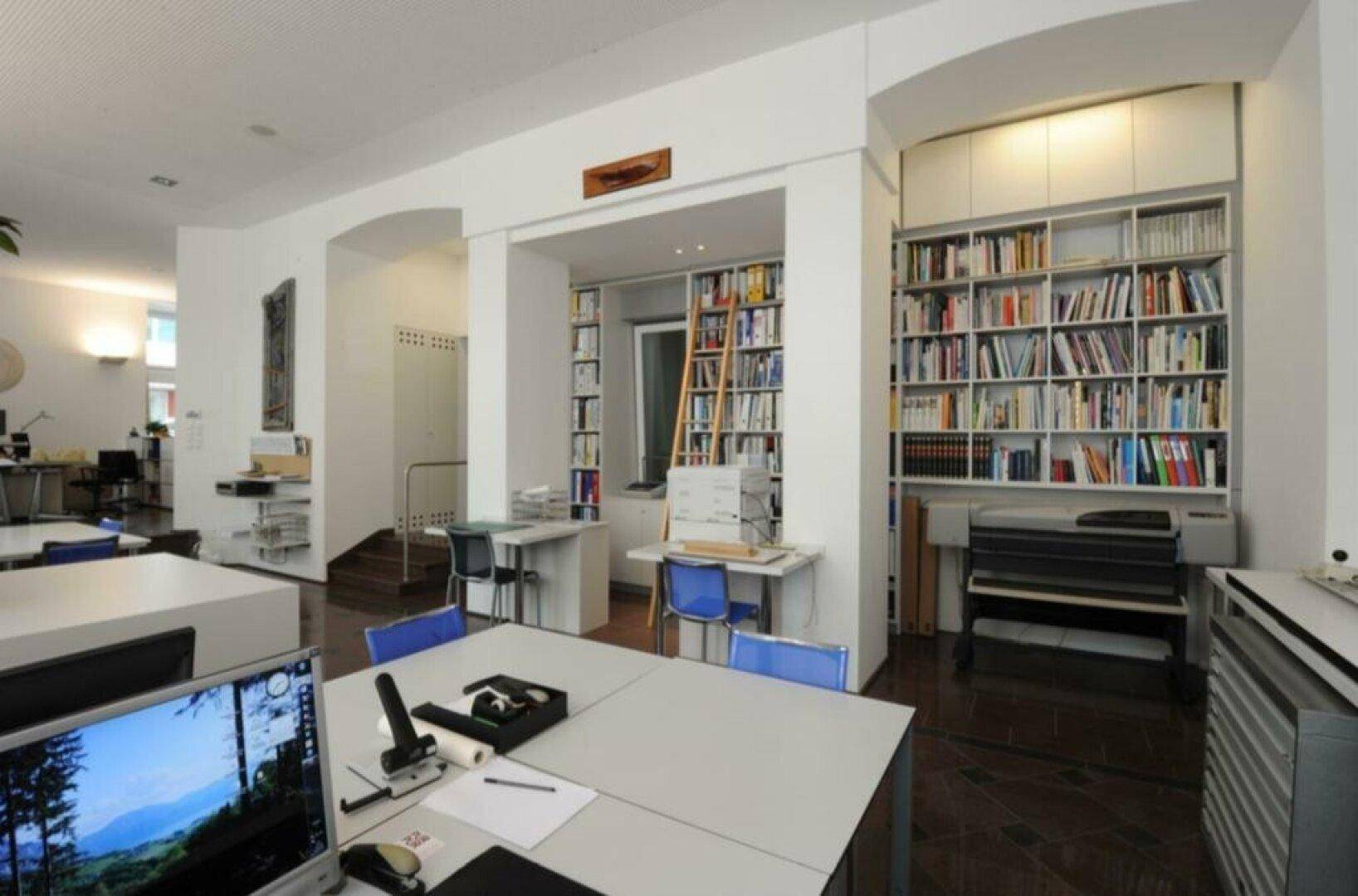 Bürobereich mit umfangreichen Bücherregalen, einer Leiter und mehreren Arbeitsplätzen.