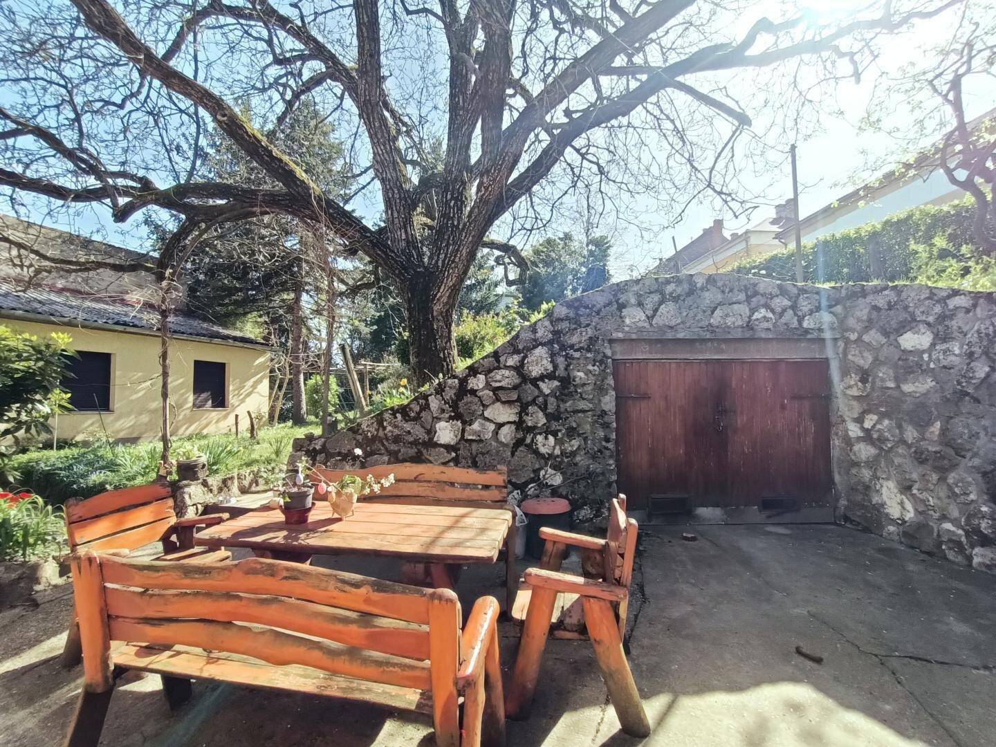Gemütlicher Essbereich im Freien mit Holztisch und Bänken neben einer Steinmauer und einem Holztor.