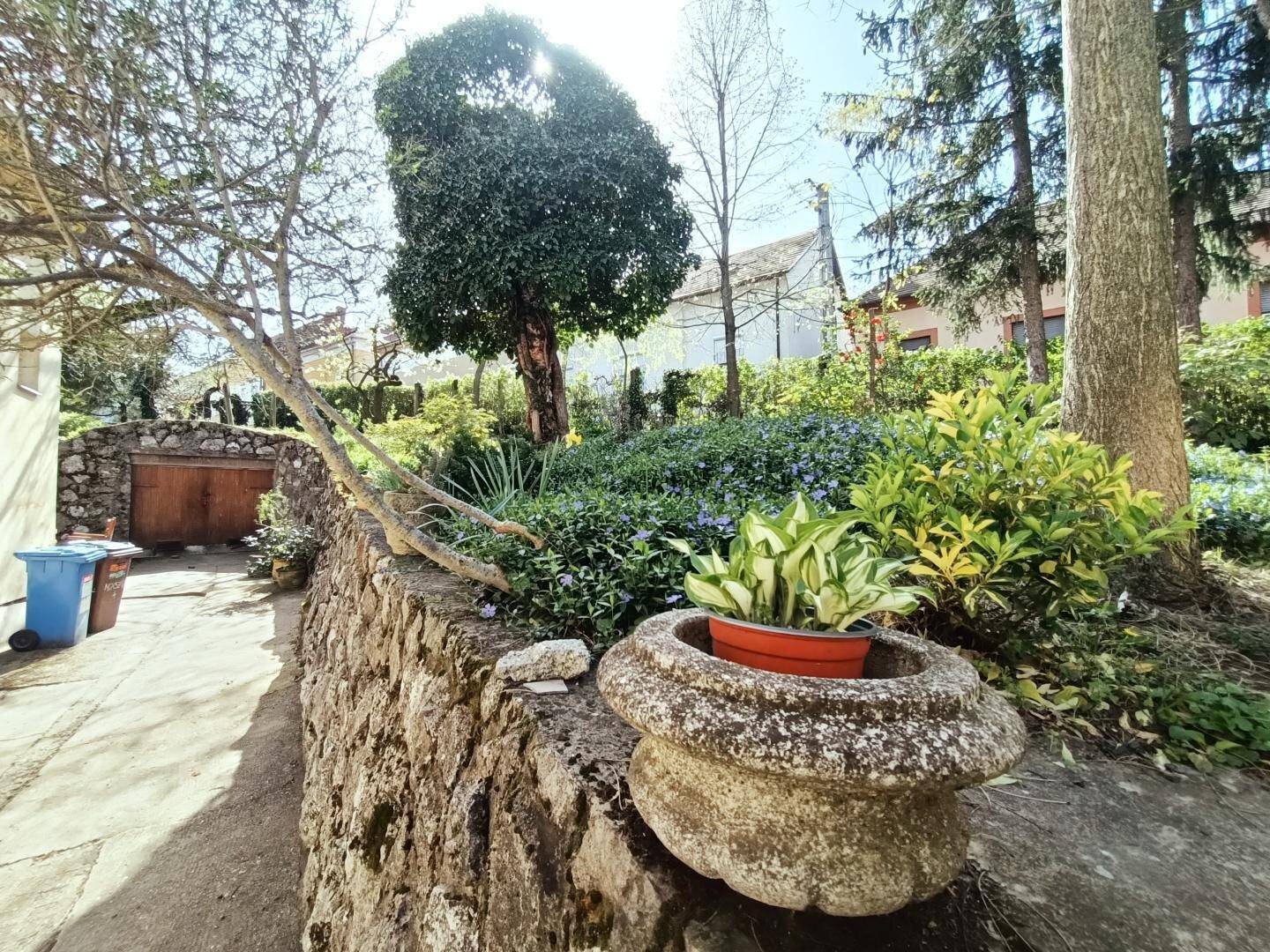 Garten mit rustikaler Steinmauer, verschiedenen Pflanzen und einem dekorativen Steintopf.