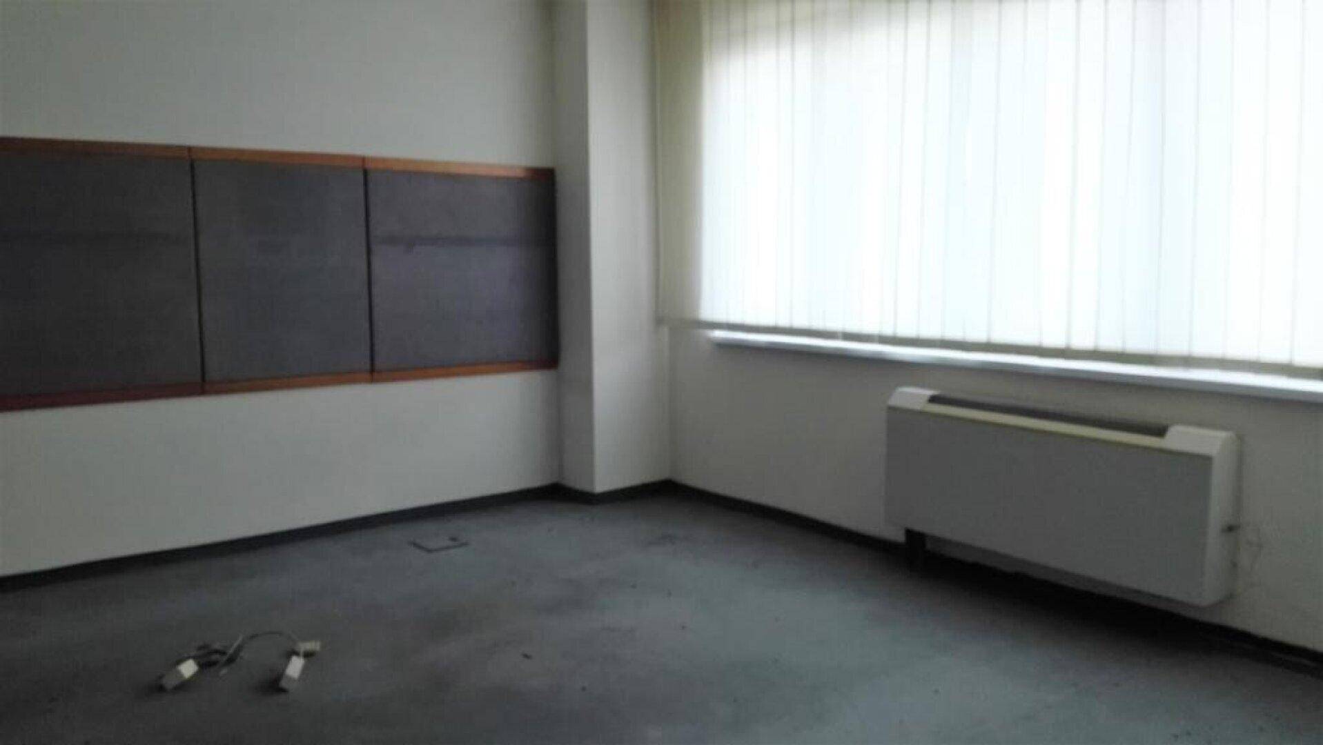 Leeres Büro mit grauem Boden, weißen Wänden, großen Fenstern und einer dunklen Tafel an der Wand.