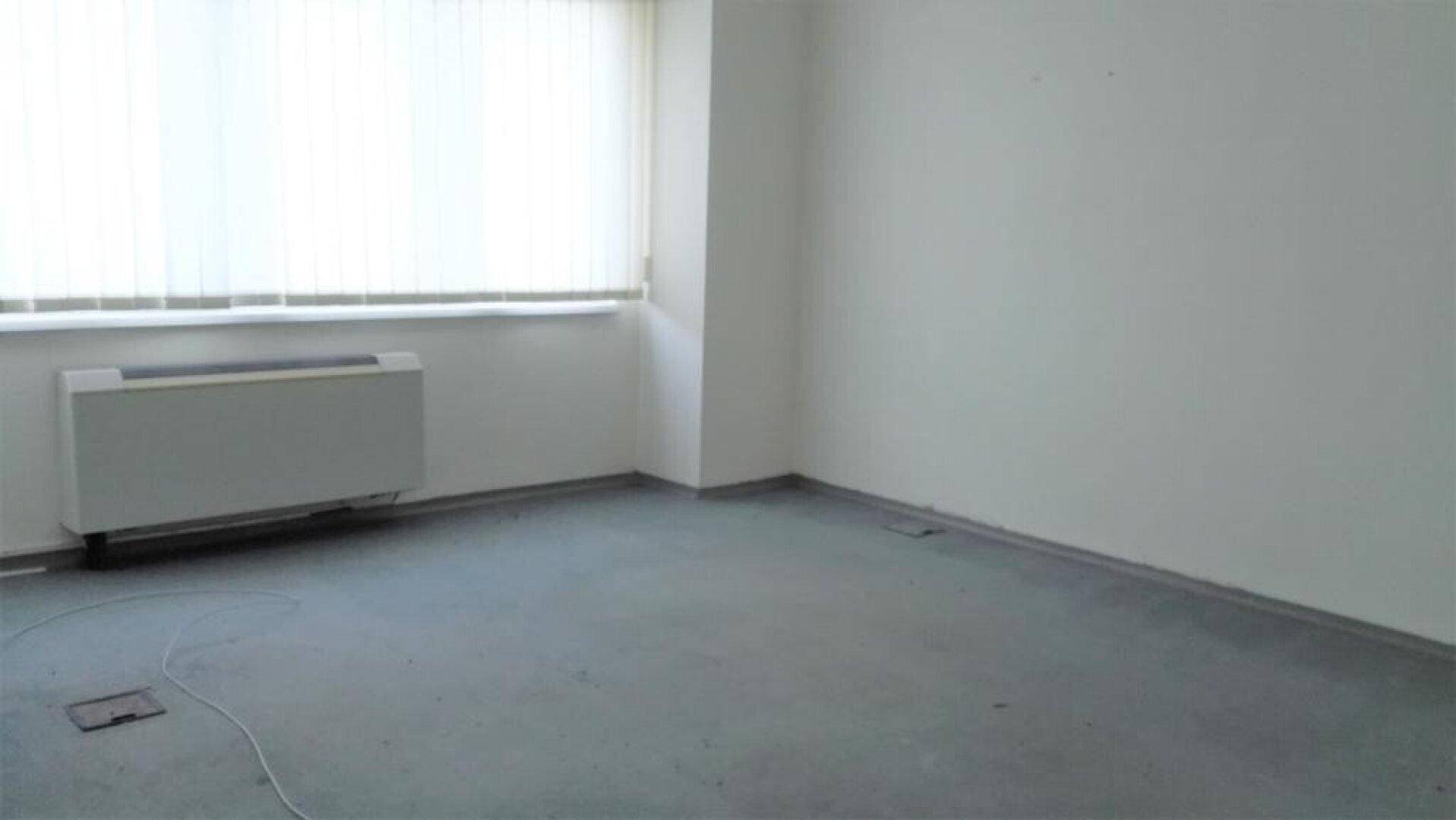 Leeres Büro mit grauem Boden, weißen Wänden, großen Fenstern mit Jalousien und einem Heizkörper.