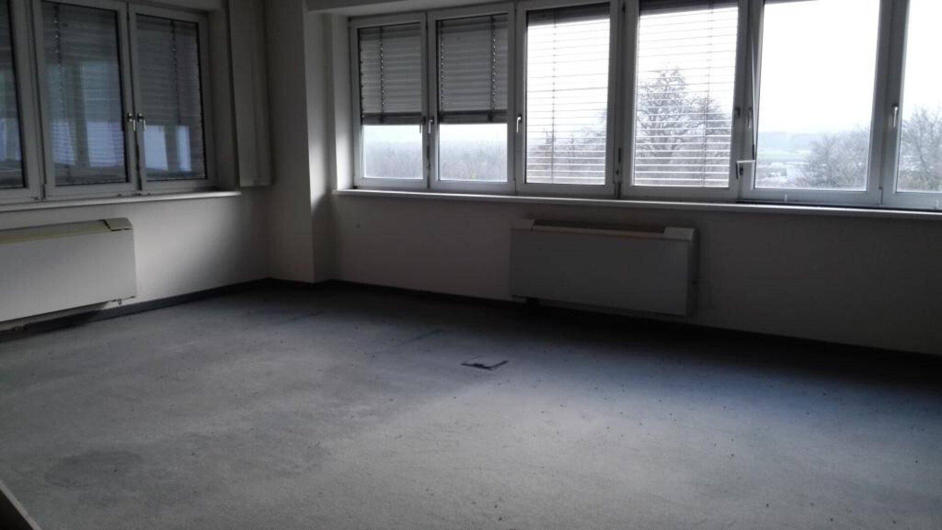 Leeres Büro mit grauem Boden, großen Fenstern mit Jalousien und einem Heizkörper.