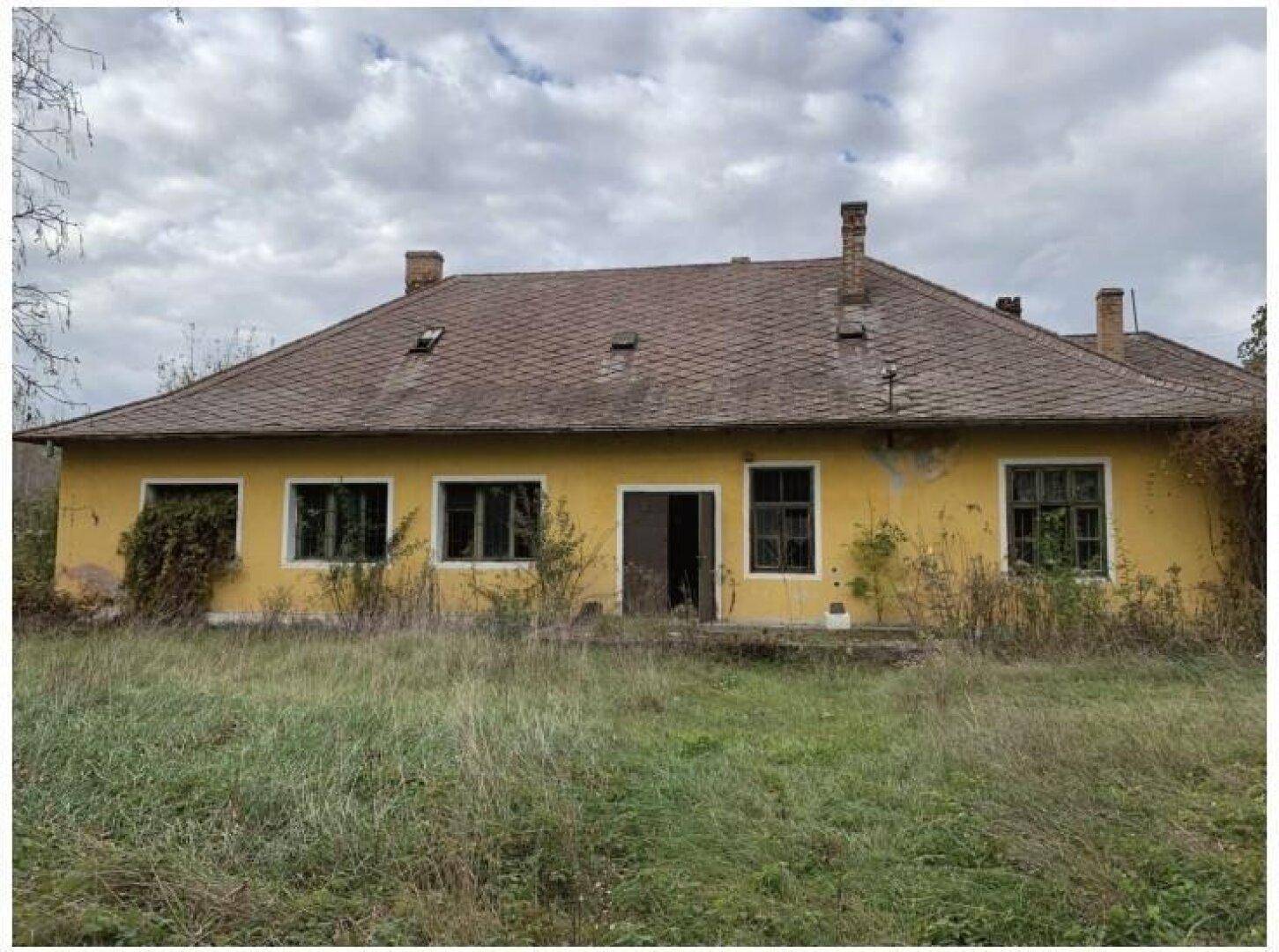Vorderansicht eines alten, gelben Hauses mit mehreren Fenstern und einem roten Ziegeldach auf einer Wiese.