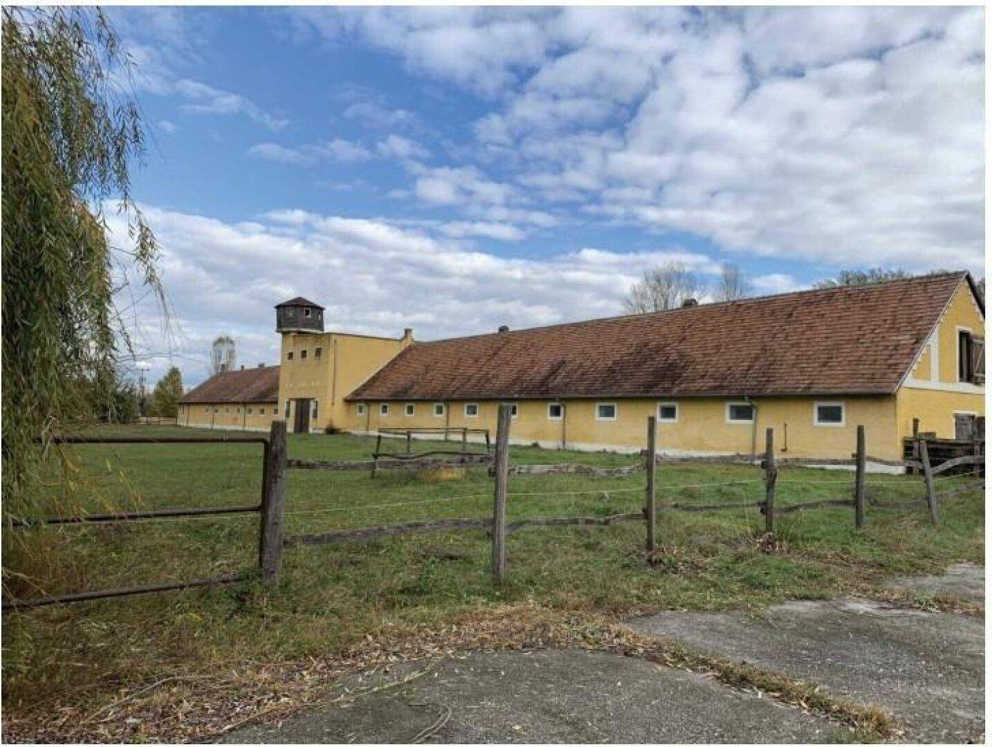 Langes, gelbes Gebäude mit rotem Ziegeldach und einem Turm, umgeben von einer Wiese und einem Holzzaun.