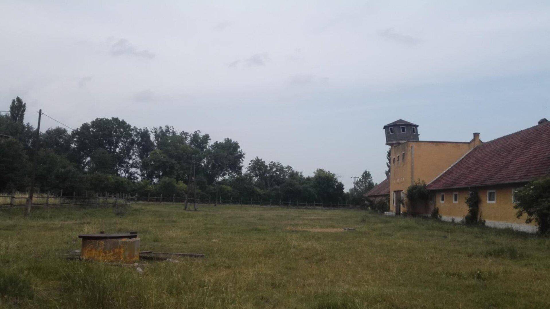 Weitläufige Wiesenfläche mit einem alten Brunnen und einem langen Gebäude im Hintergrund unter bewölktem Himmel.