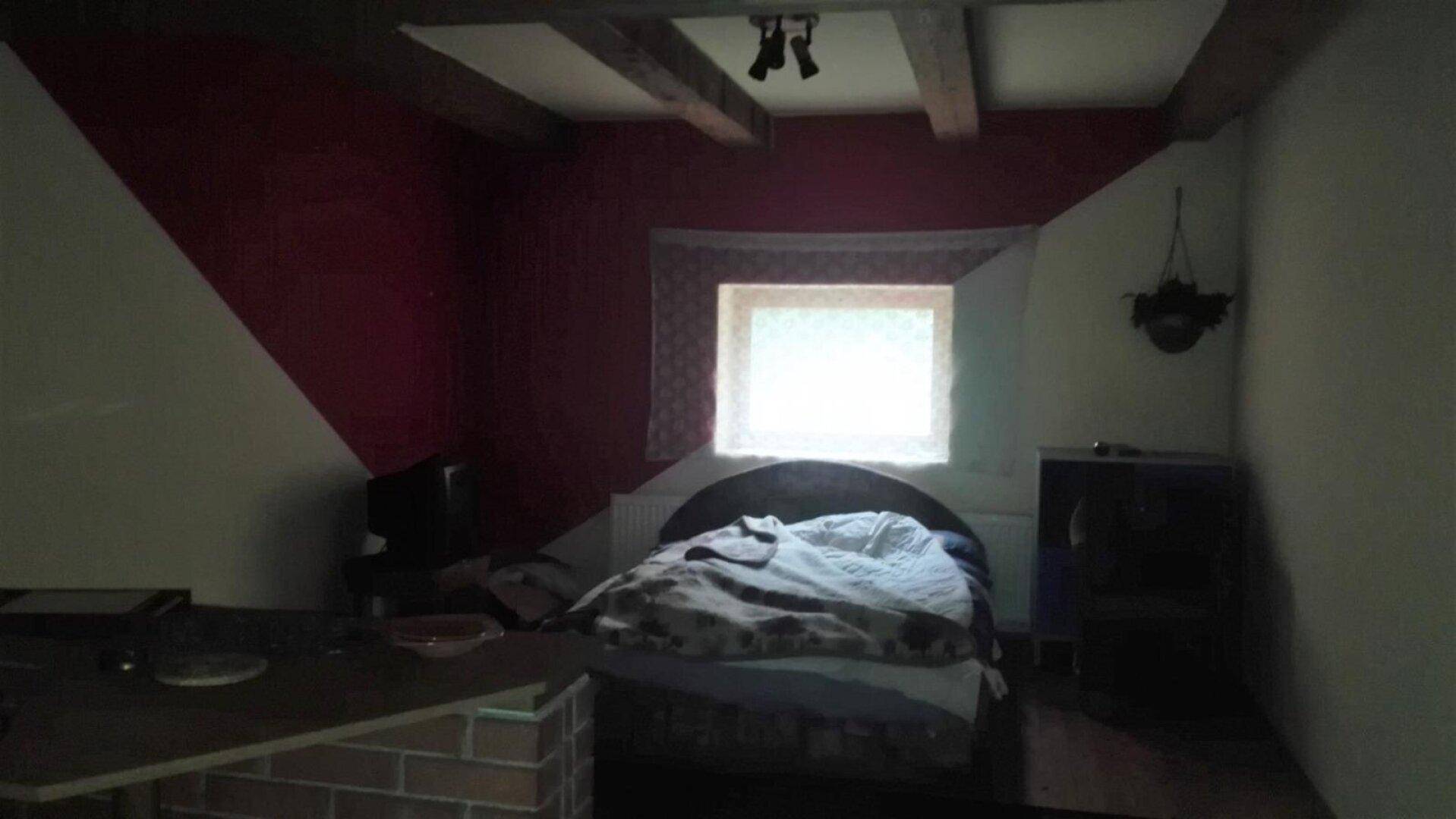 Schlafzimmer mit schrägen Wänden, einem Bett, einem kleinen Fenster und sichtbaren Holzbalken an der Decke.