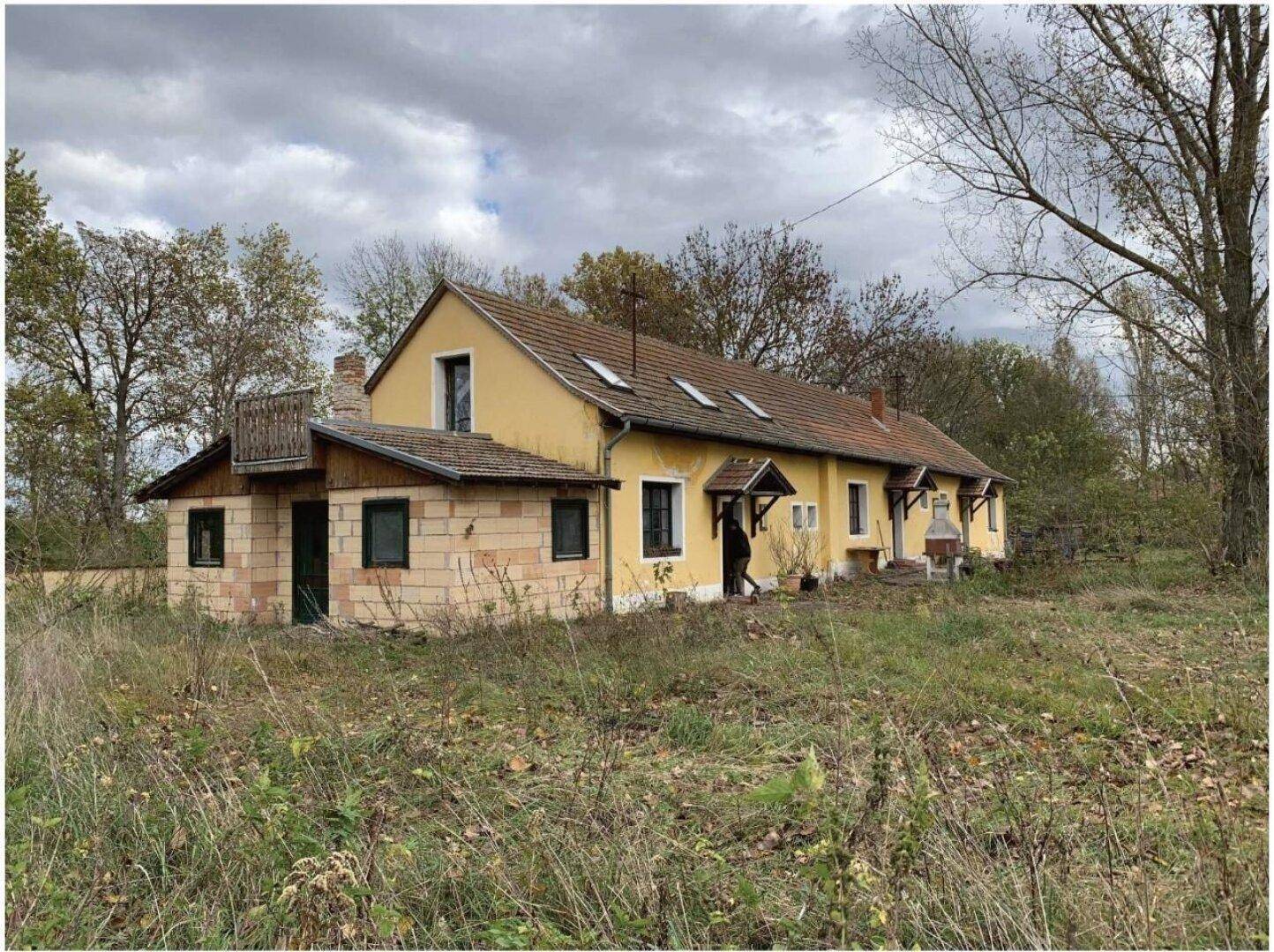 Langes, gelbes Gebäude mit rotem Ziegeldach und einem Anbau aus hellen Ziegeln, umgeben von Natur.