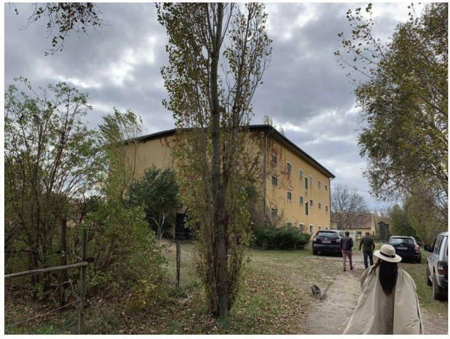 Großes, gelbes Gebäude mit vielen Fenstern, umgeben von Bäumen und parkenden Autos auf einem Feldweg.