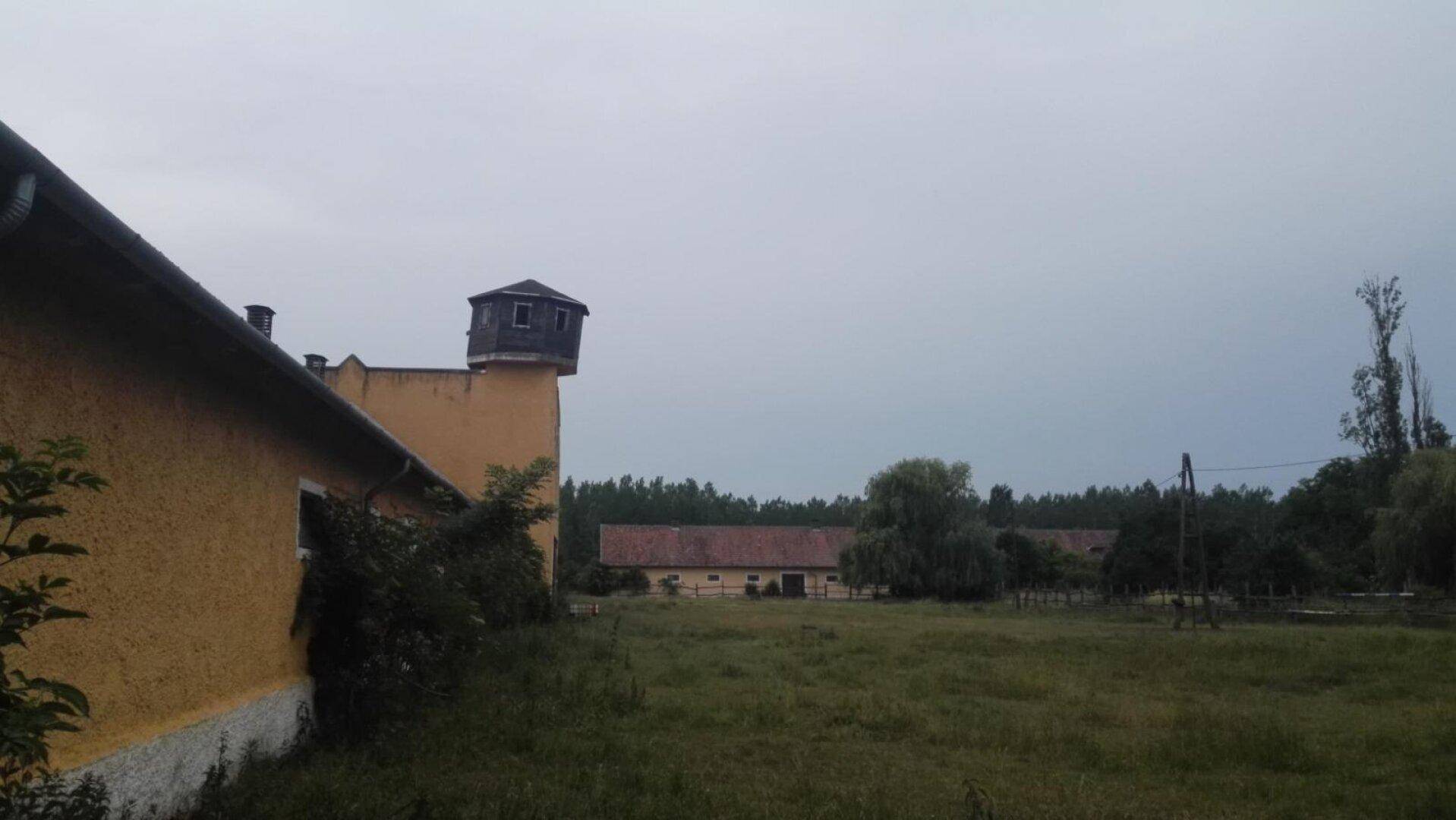 Weitläufige Landschaft mit einem gelben Gebäude und einem Turm im Hintergrund unter bewölktem Himmel.