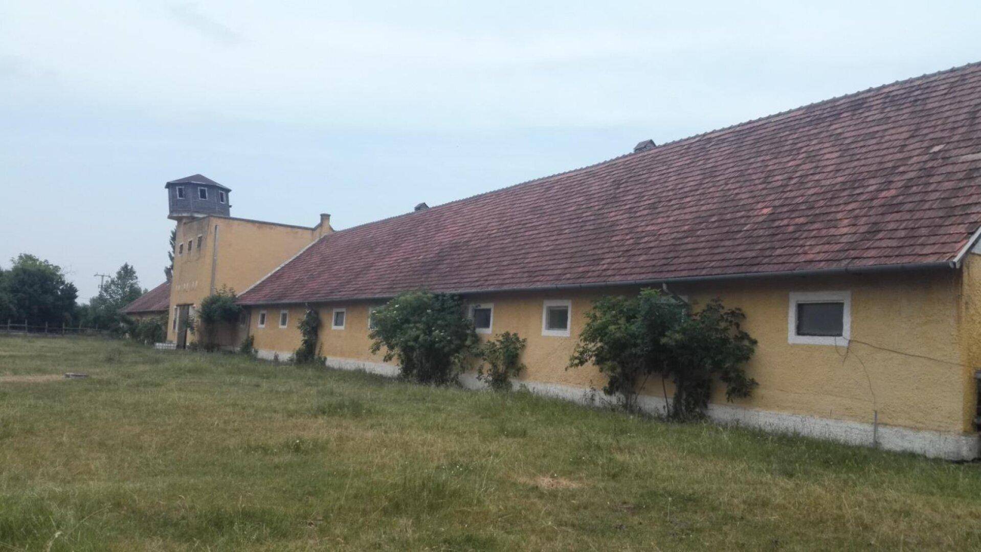 Langes, gelbes Gebäude mit rotem Ziegeldach und einem kleinen Turm, umgeben von einer weiten Wiese.
