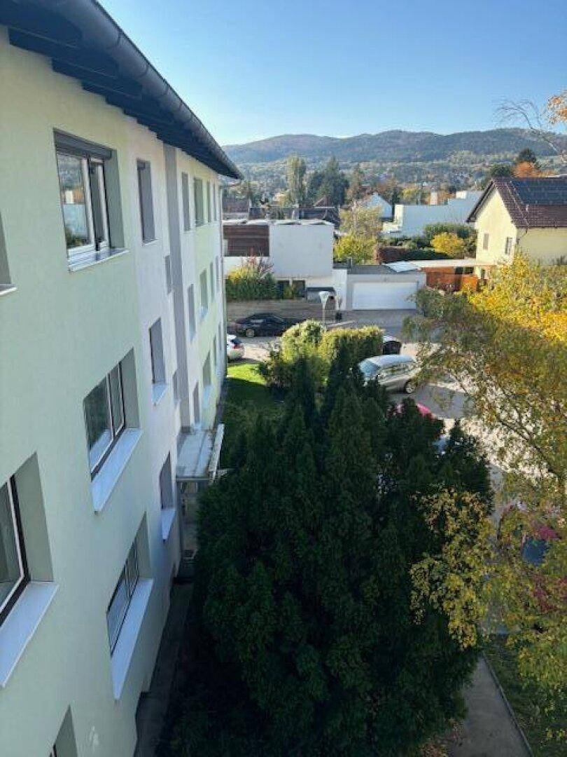 Blick vom Balkon auf die Umgebung mit Bäumen und Bergen im Hintergrund.
