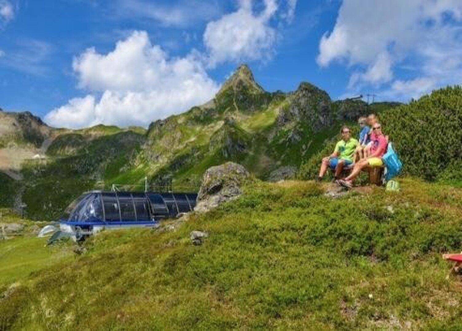 Wanderer entspannen sich auf einem grünen Hügel mit Blick auf die Bergwelt und eine Seilbahnstation.