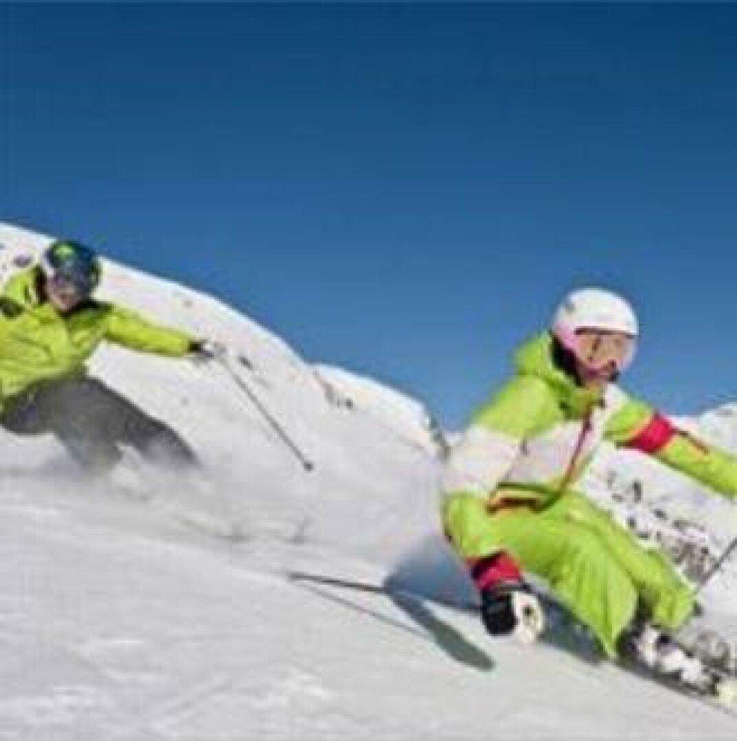 Zwei Skifahrer genießen die Abfahrt auf einer verschneiten Piste mit strahlendem Sonnenschein.