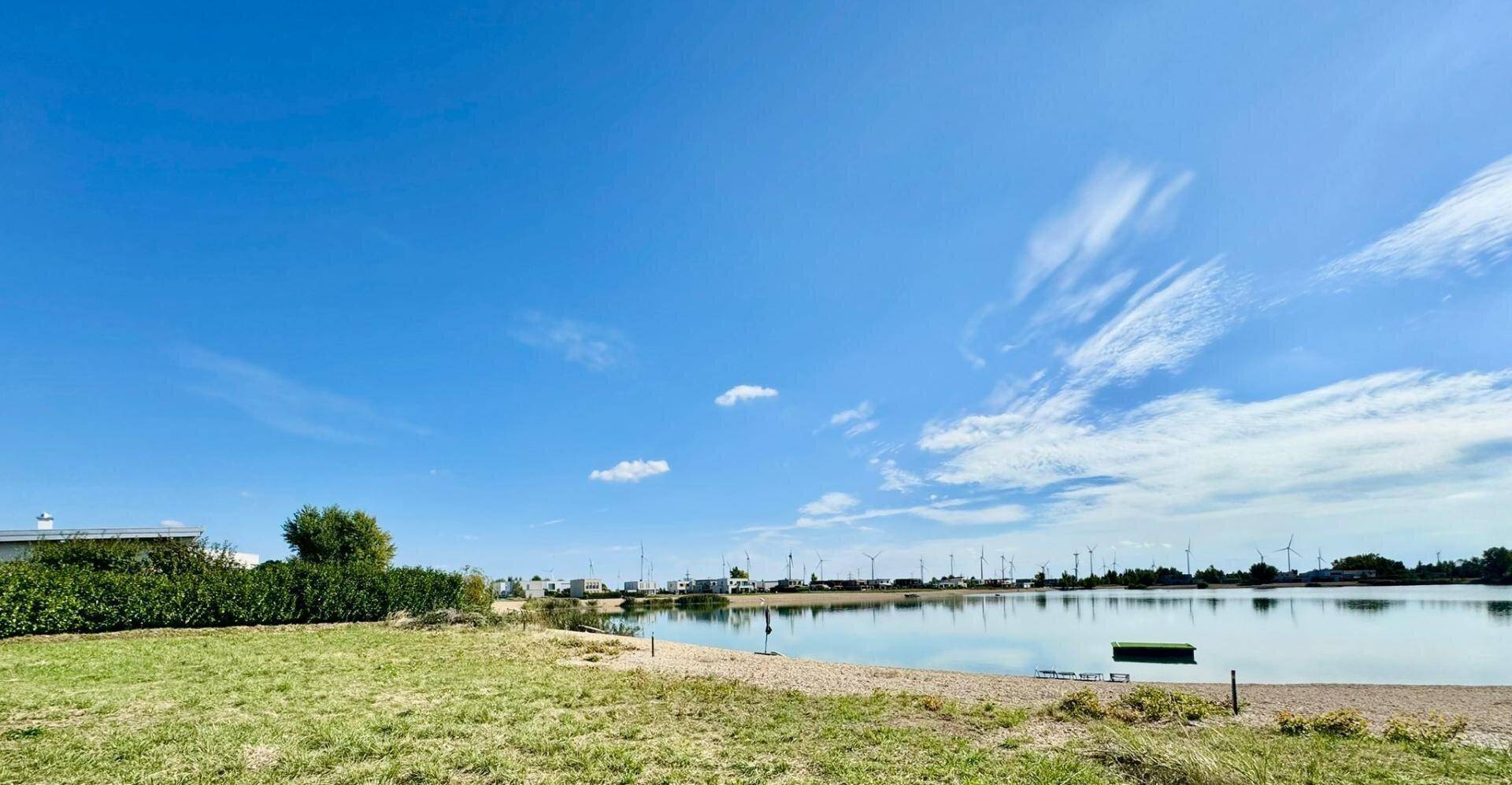 Panoramablick auf den See mit Sandufer, umgeben von grünen Flächen und modernen Gebäuden in der Ferne.