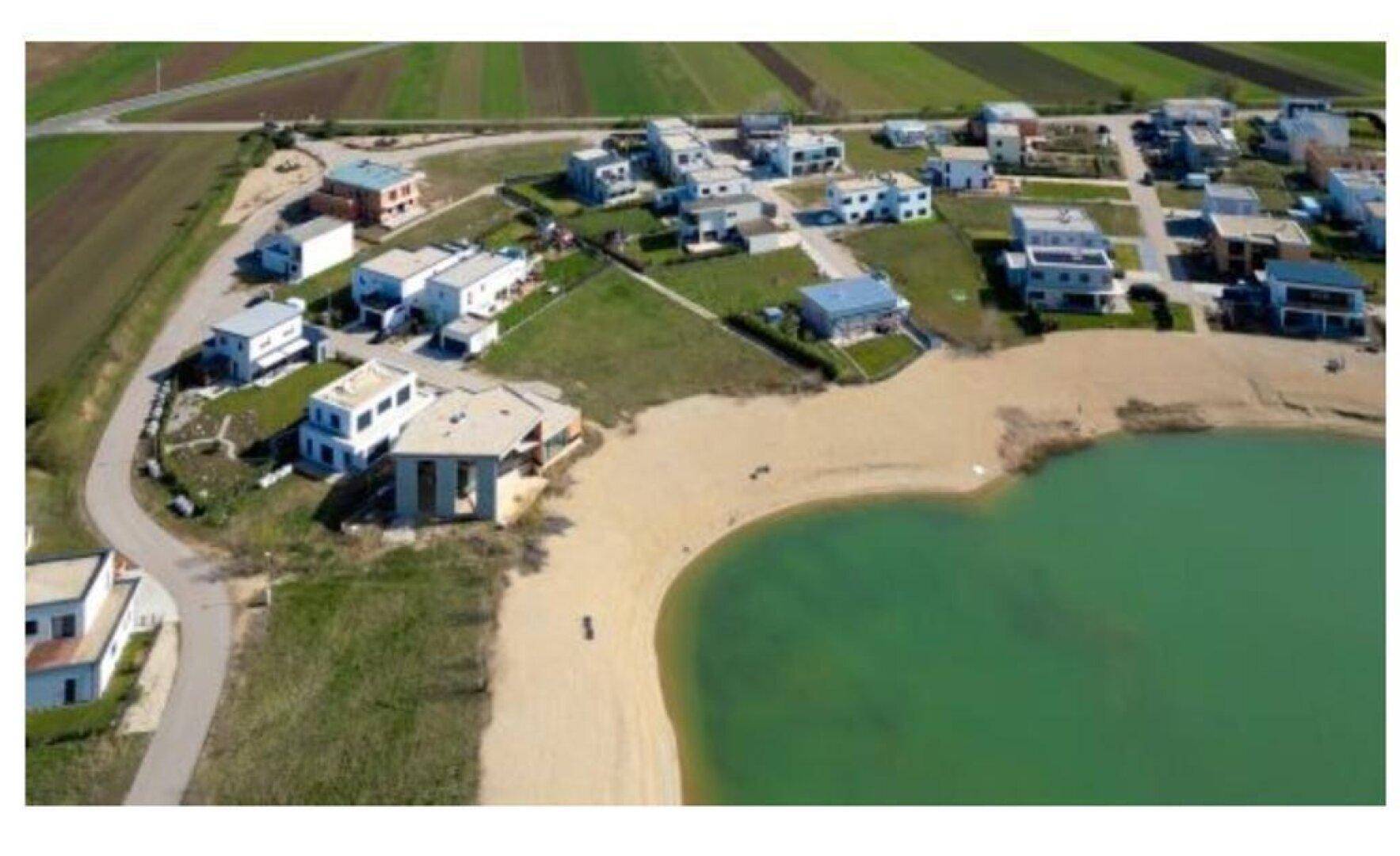 Luftaufnahme einer modernen Wohnsiedlung am Seeufer mit privatem Sandstrand und umliegenden Feldern.
