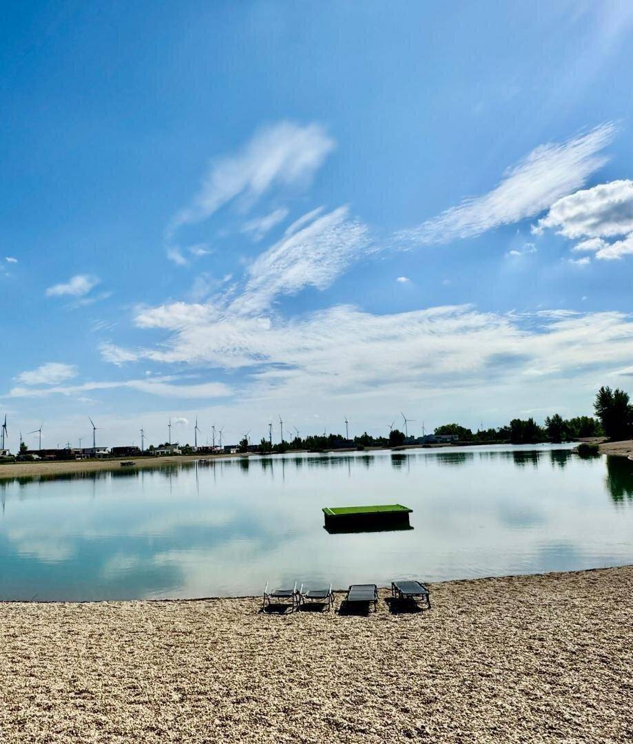 Idyllischer Sandstrand am See mit Sonnenliegen und einer schwimmenden Plattform unter blauem Himmel.