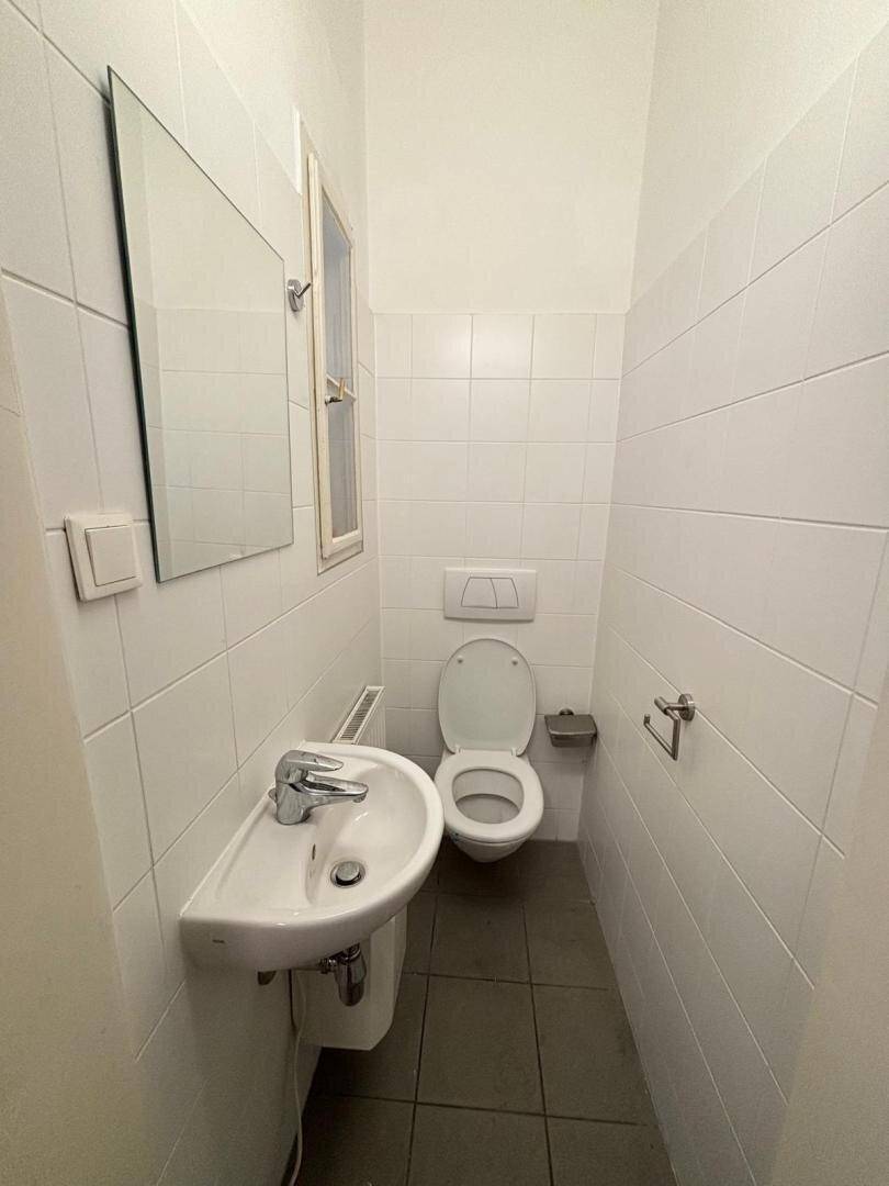 Schmale Gästetoilette mit weißen Fliesen, Waschbecken, WC und einem kleinen Fenster.