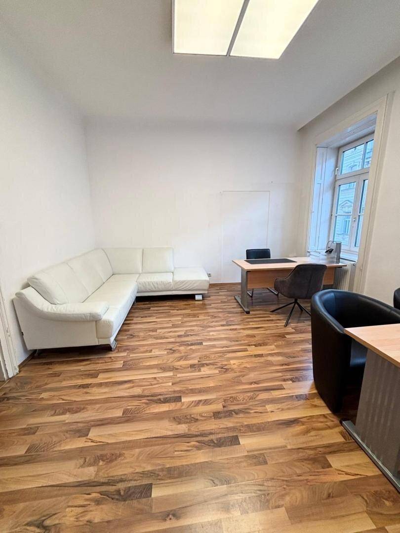 Geräumiges Büro mit Holzboden, weißem Ecksofa und einem Schreibtisch am Fenster.