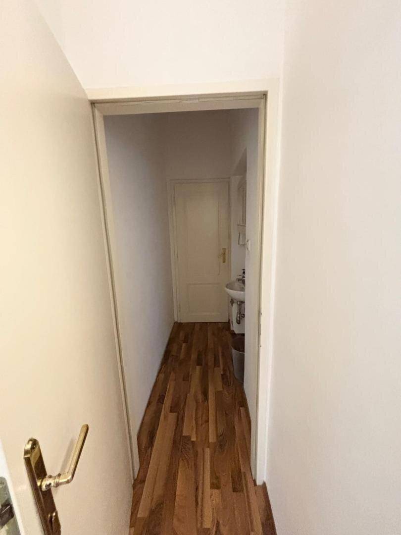 Schmaler Flur mit Holzboden, weißen Wänden und einer Tür am Ende, die zu einem Bad führt.