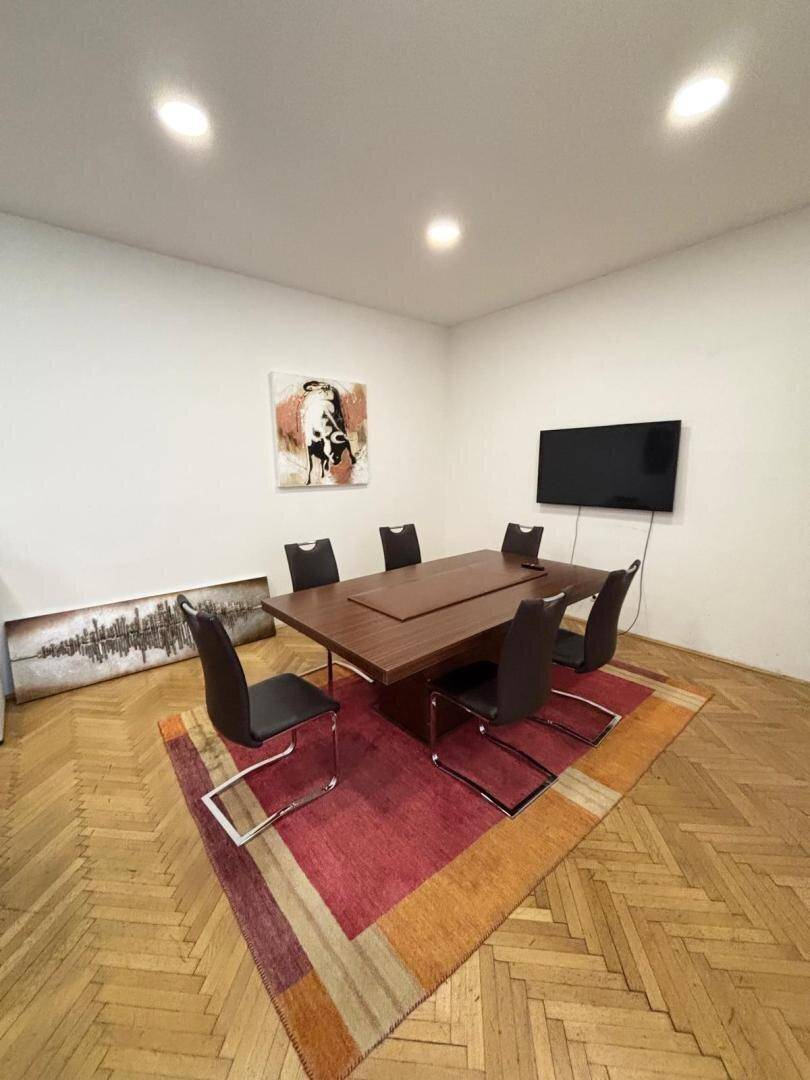 Konferenzraum mit Holzboden, großem Tisch, Stühlen und einem Wandfernseher.