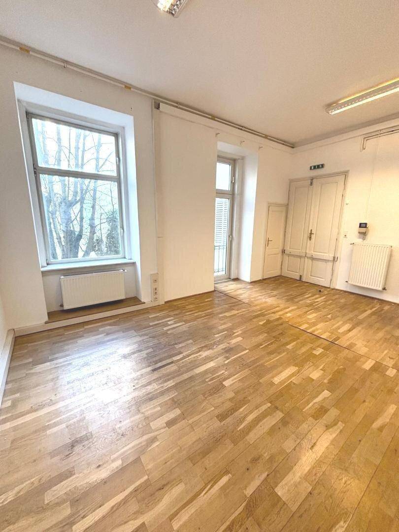 Geräumiges, leeres Büro mit Holzboden, großen Fenstern und viel Tageslicht.