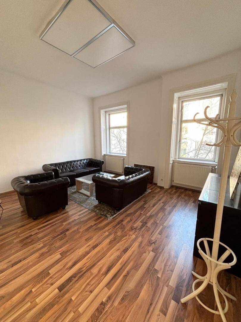 Wohnzimmer mit Holzboden, zwei großen Fenstern und schwarzen Ledersofas.