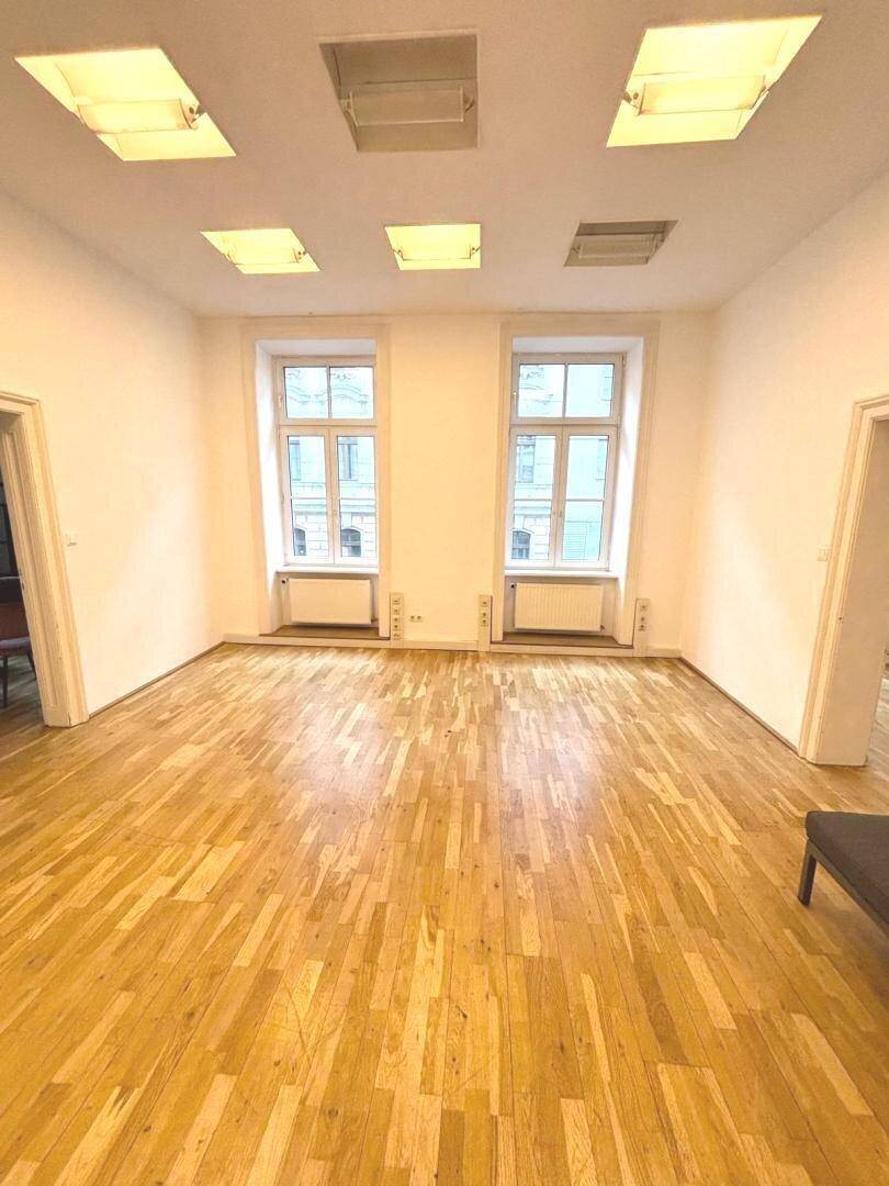 Helles, leeres Büro mit Holzboden, zwei großen Fenstern und Deckenbeleuchtung.
