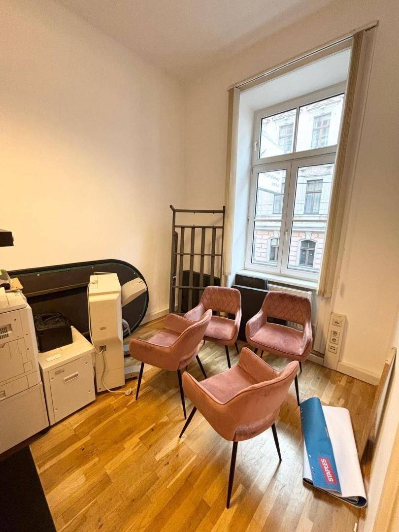 Kleiner Büroraum mit Holzboden, einem großen Fenster und vier rosa Stühlen.
