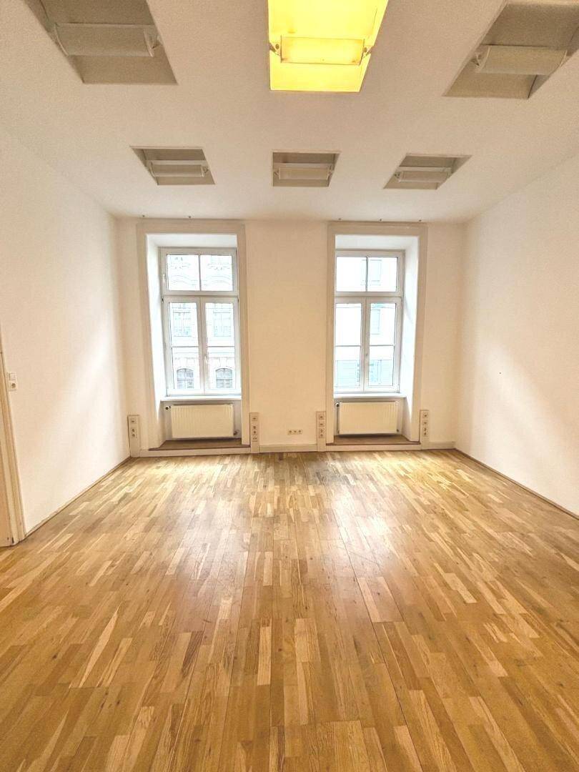 Helles, leeres Büro mit Holzboden, zwei großen Fenstern und Deckenbeleuchtung.