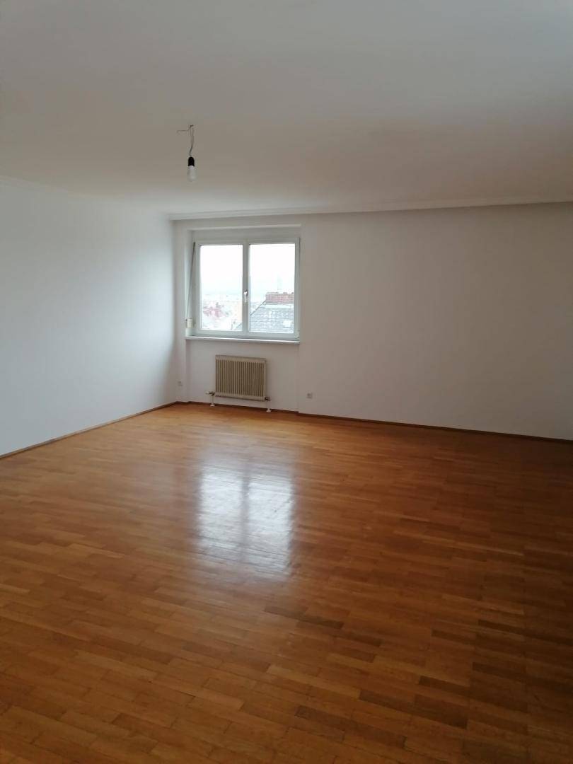 Geräumiges, leeres Wohnzimmer mit hellem Parkettboden und einem großen Fenster mit Stadtblick.