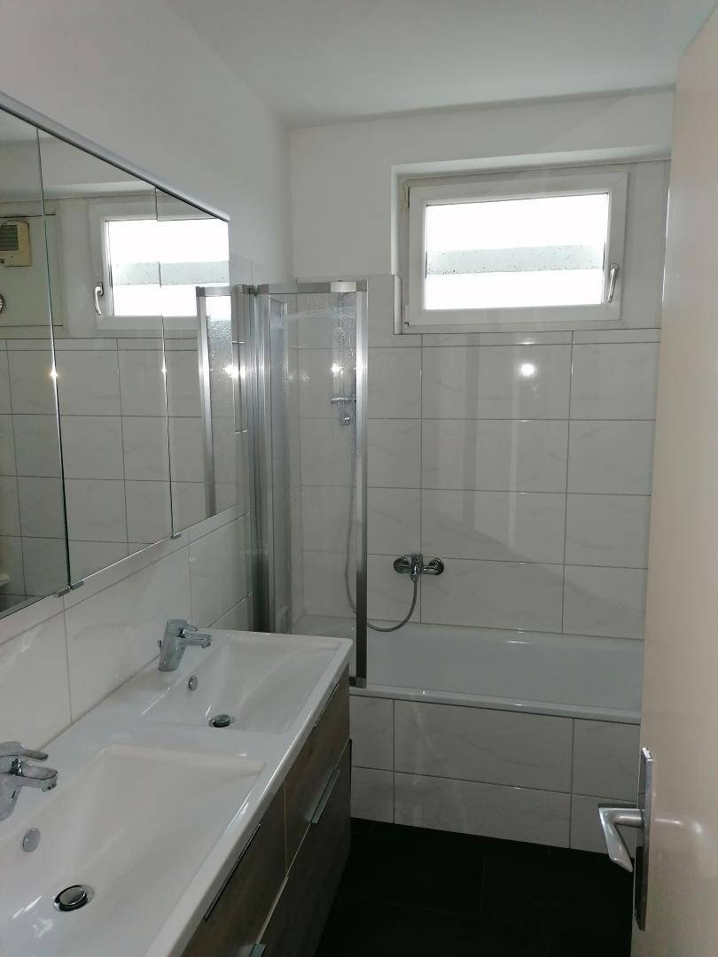 Modernes Badezimmer mit Doppelwaschbecken, Badewanne und Duschabtrennung sowie zwei Fenstern.