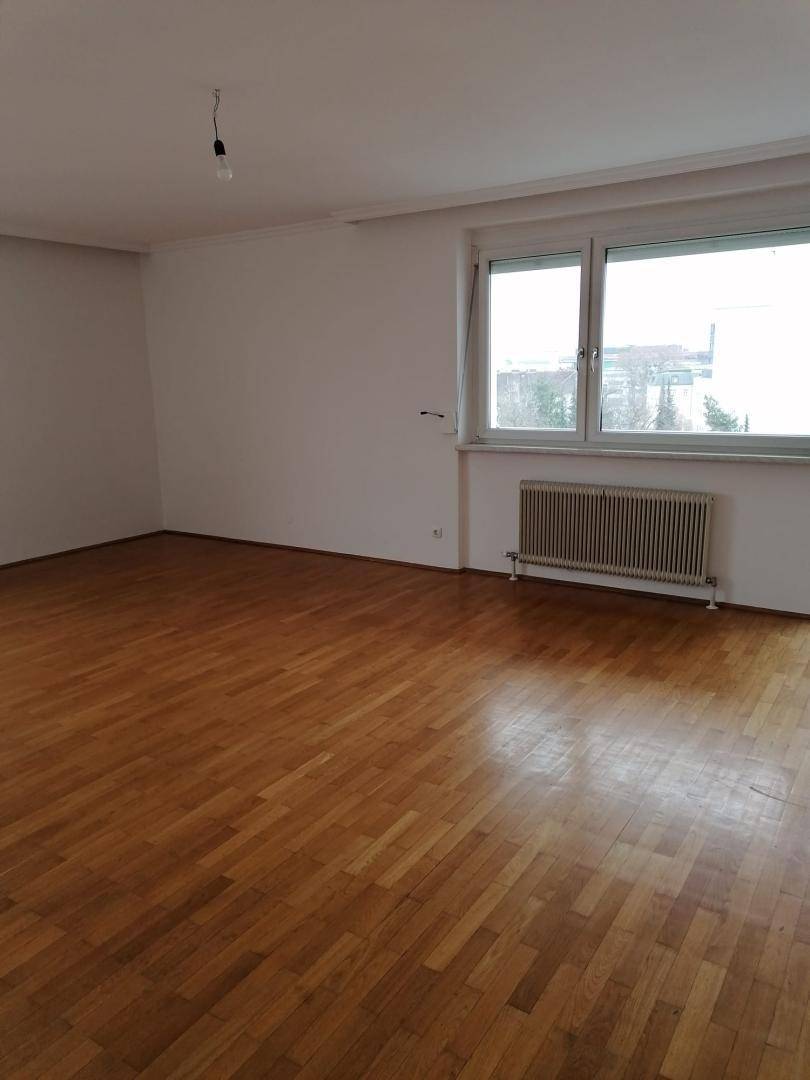 Geräumiges, leeres Wohnzimmer mit hellem Parkettboden und einem großen Fenster mit Stadtblick.