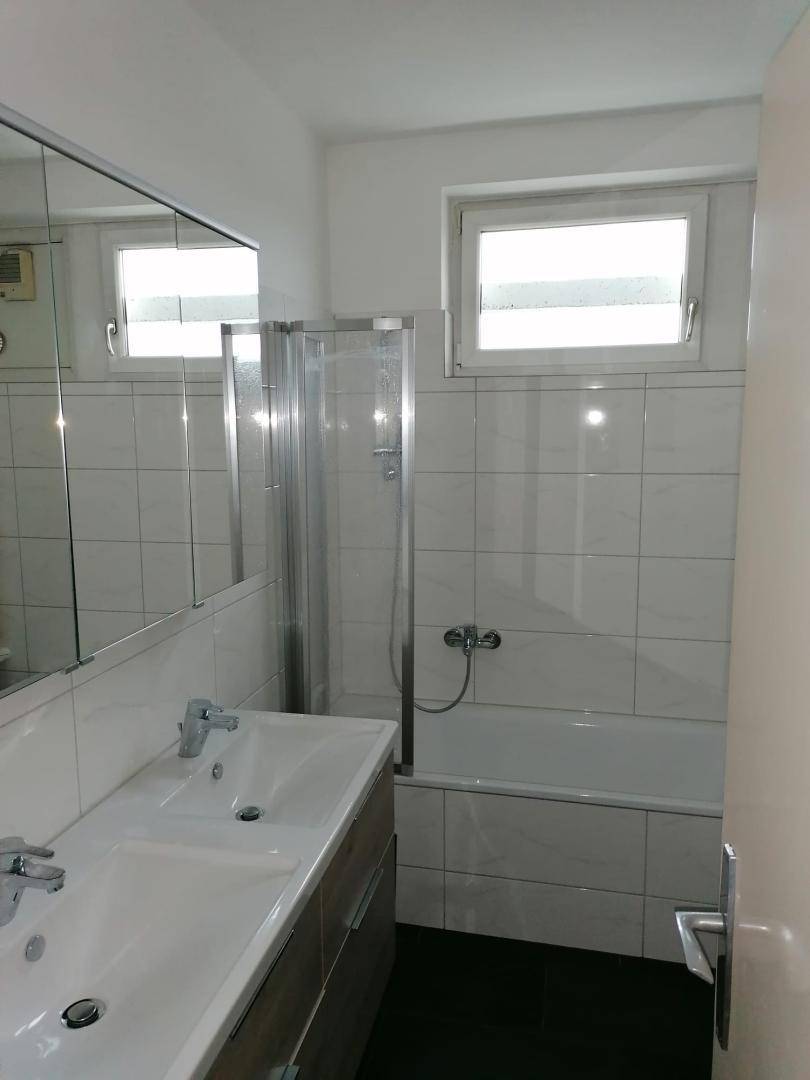 Modernes Badezimmer mit Doppelwaschbecken, Badewanne und Duschabtrennung sowie zwei Fenstern.