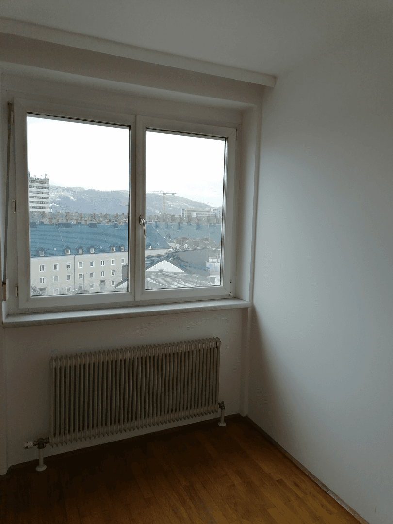Helles Schlafzimmer mit Holzboden, Heizkörper und einem Fenster mit Blick auf die Stadt.