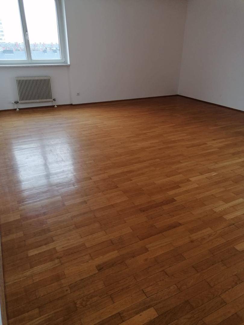 Geräumiges, leeres Wohnzimmer mit hellem Parkettboden und einem großen Fenster mit Stadtblick.