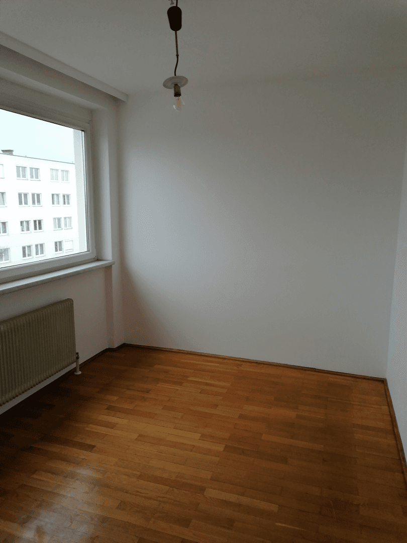 Helles Zimmer mit Holzboden und einem Fenster, das einen Blick auf die umliegenden Gebäude bietet.