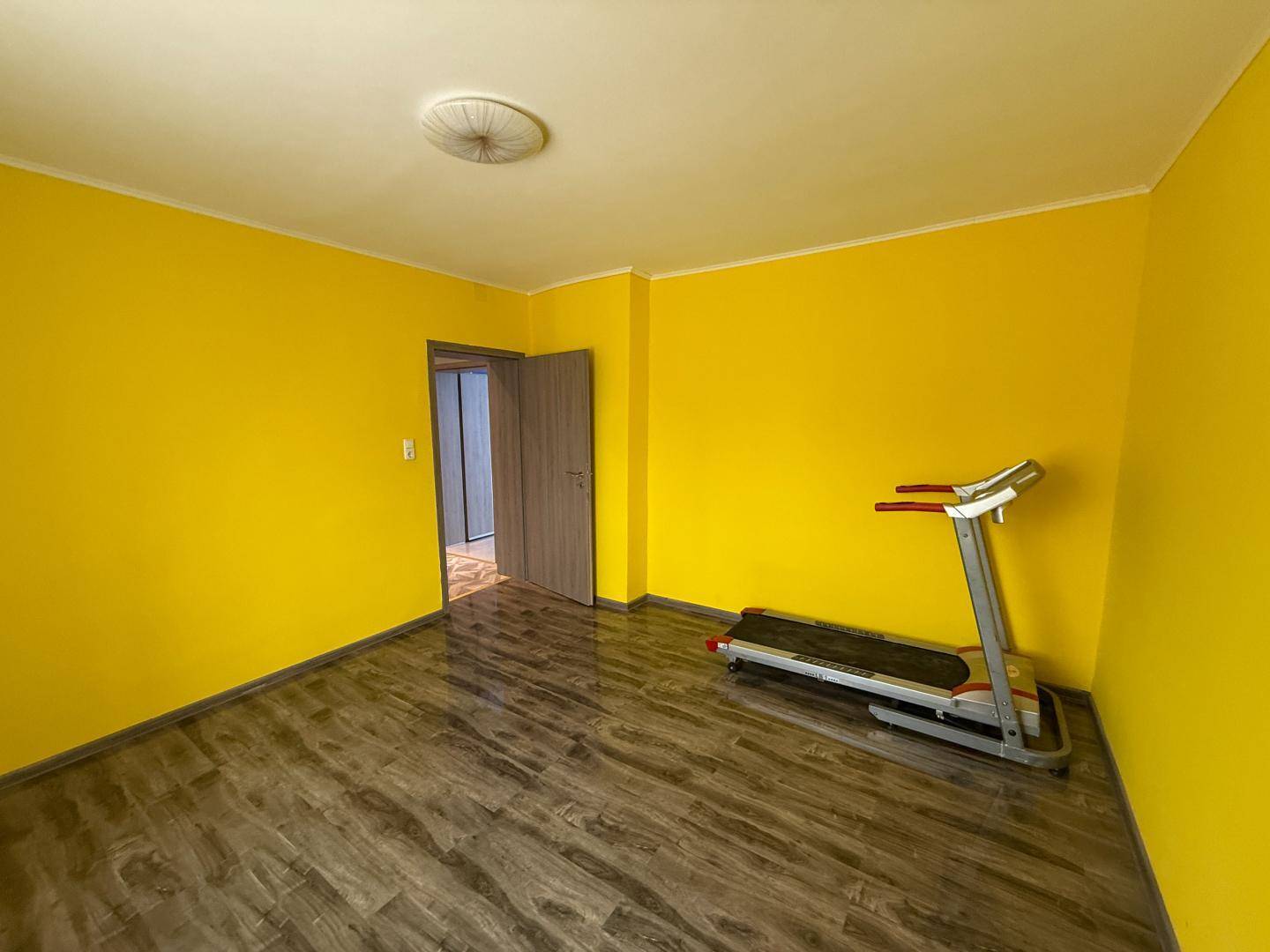 Helles Zimmer mit gelben Wänden, Laminatboden und Laufband.