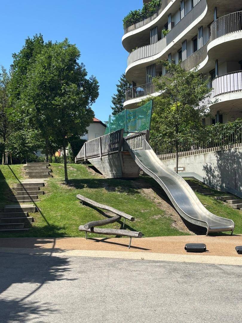 Große Rutsche auf einem Spielplatz mit Bäumen und einem modernen Gebäude im Hintergrund.