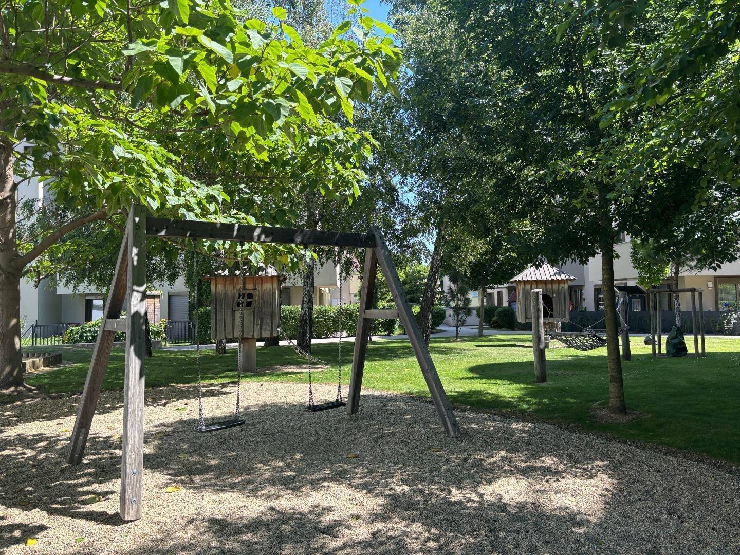 Spielplatz mit Schaukeln und Bäumen, umgeben von einer grünen Wohnanlage.