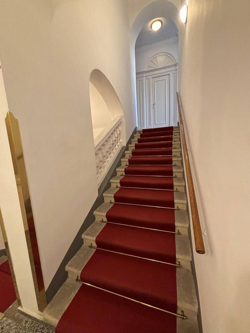 Elegantes Treppenhaus mit rotem Teppichläufer, weißem Geländer und einer klassischen Tür am oberen Ende.