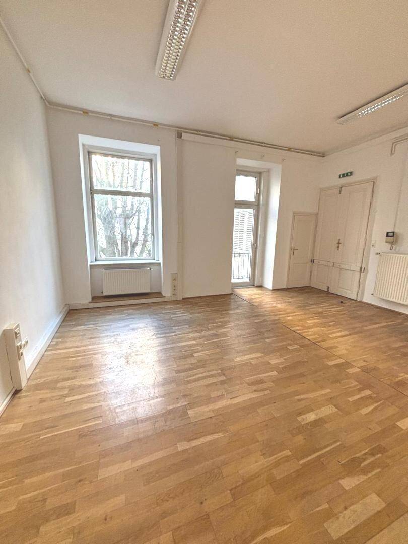 Geräumiger Raum mit Holzboden, großem Fenster und Zugang zu einem kleinen Balkon, geeignet für Bürozwecke.