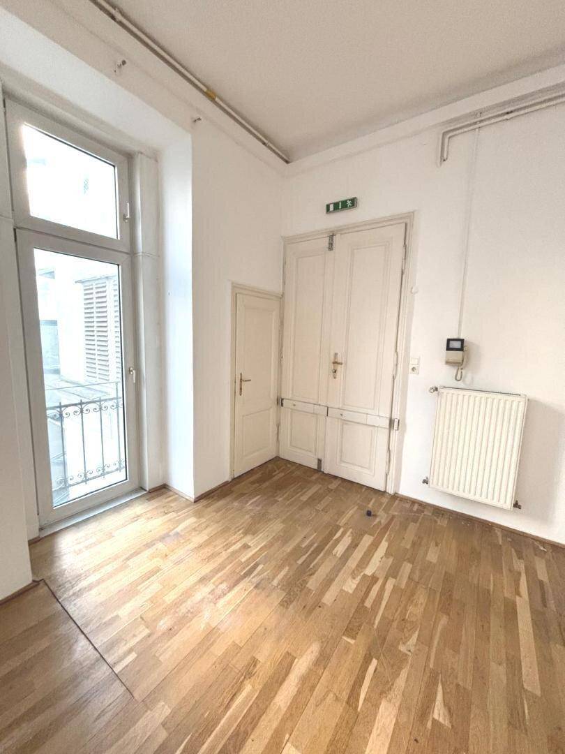 Ecke eines Raumes mit Holzboden, einer Tür zum Balkon und einem Heizkörper unter dem Fenster.