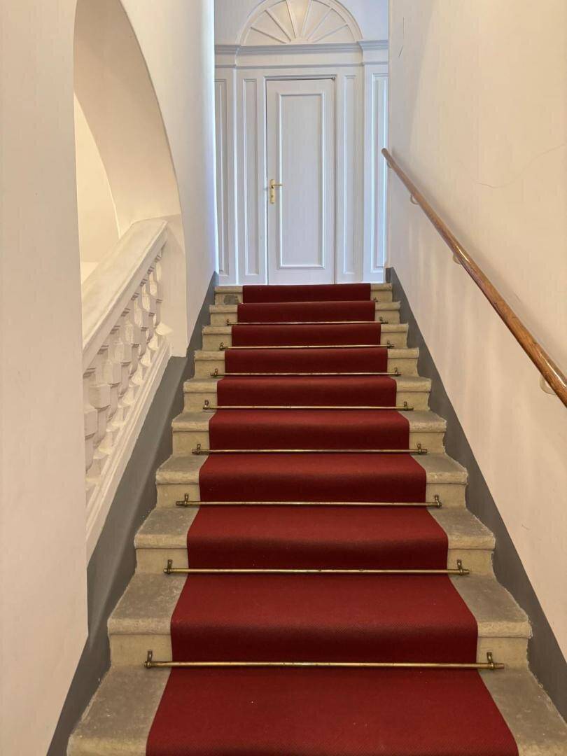 Elegantes Treppenhaus mit rotem Teppichläufer, weißem Geländer und einer klassischen Tür am oberen Ende.
