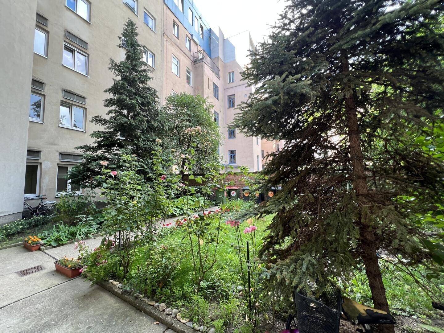 Grüner Innenhof mit hohen Bäumen, blühenden Pflanzen und Blick auf die umliegenden Gebäude.