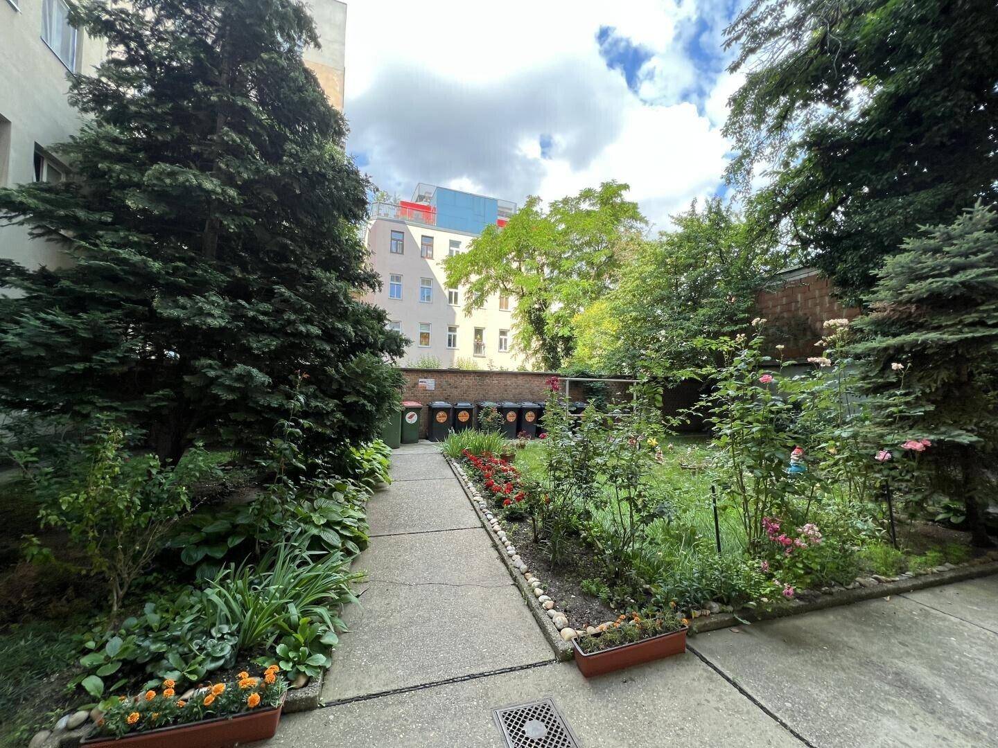 Gepflegter Gemeinschaftsgarten mit Blumenbeeten, Bäumen und einem gepflasterten Weg.