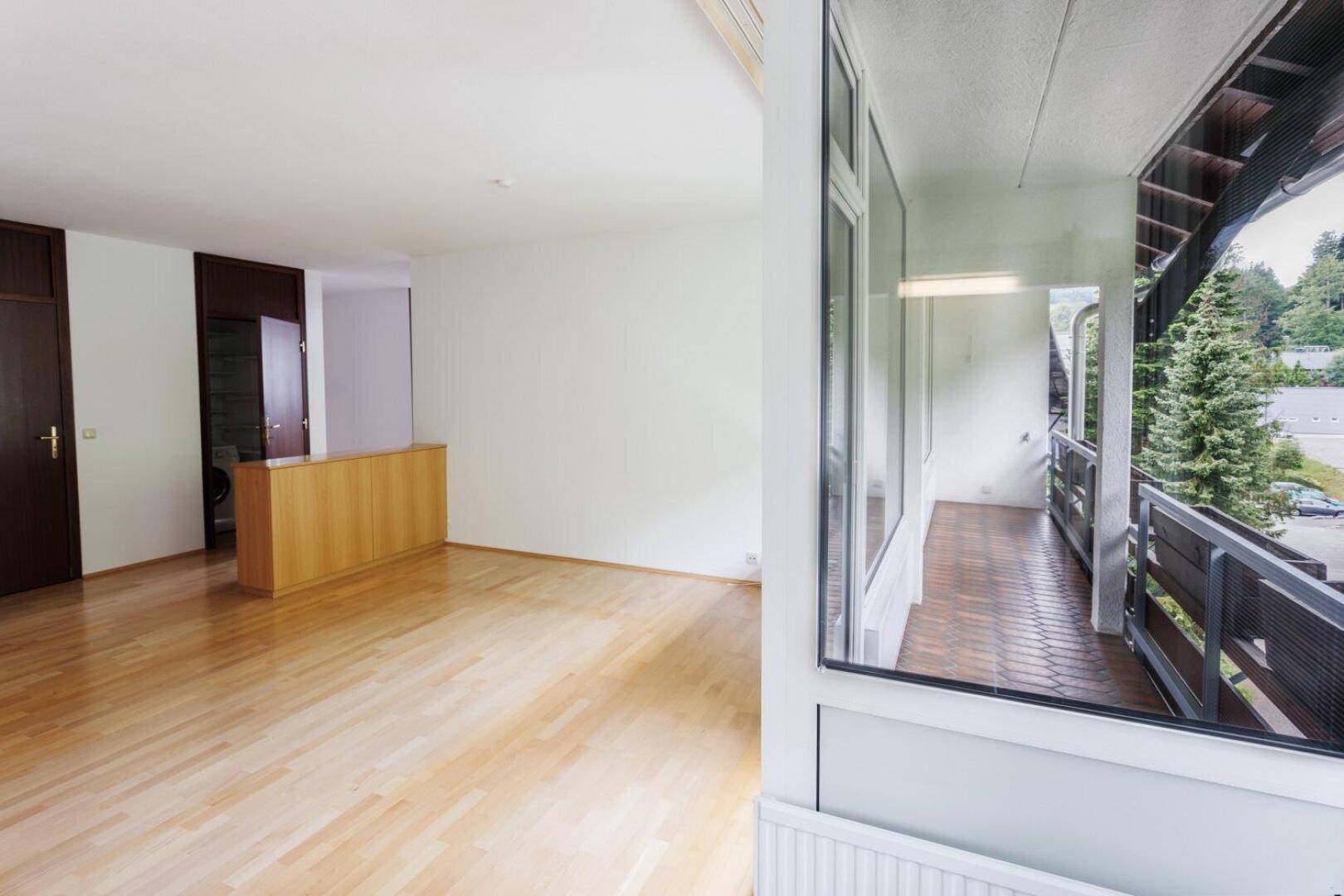 Heller Innenraum mit Holzparkett und Blick auf einen überdachten Balkon mit Fliesenboden.