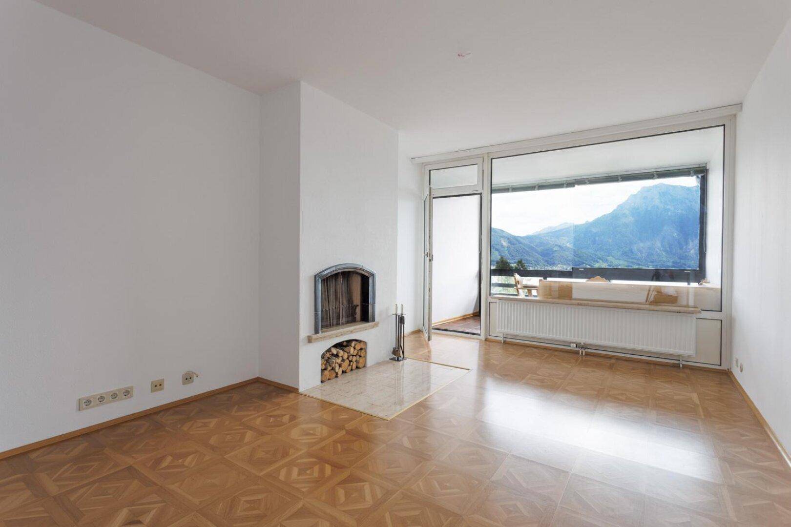 Wohnzimmer mit Holzboden, Kamin und großen Fenstern mit Blick auf Berge und See.
