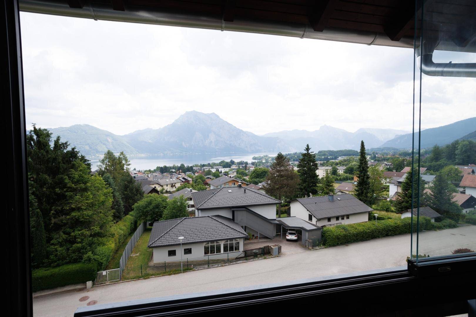 Blick auf den See und die Berge durch ein Fenster, umgeben von Häusern und Natur.