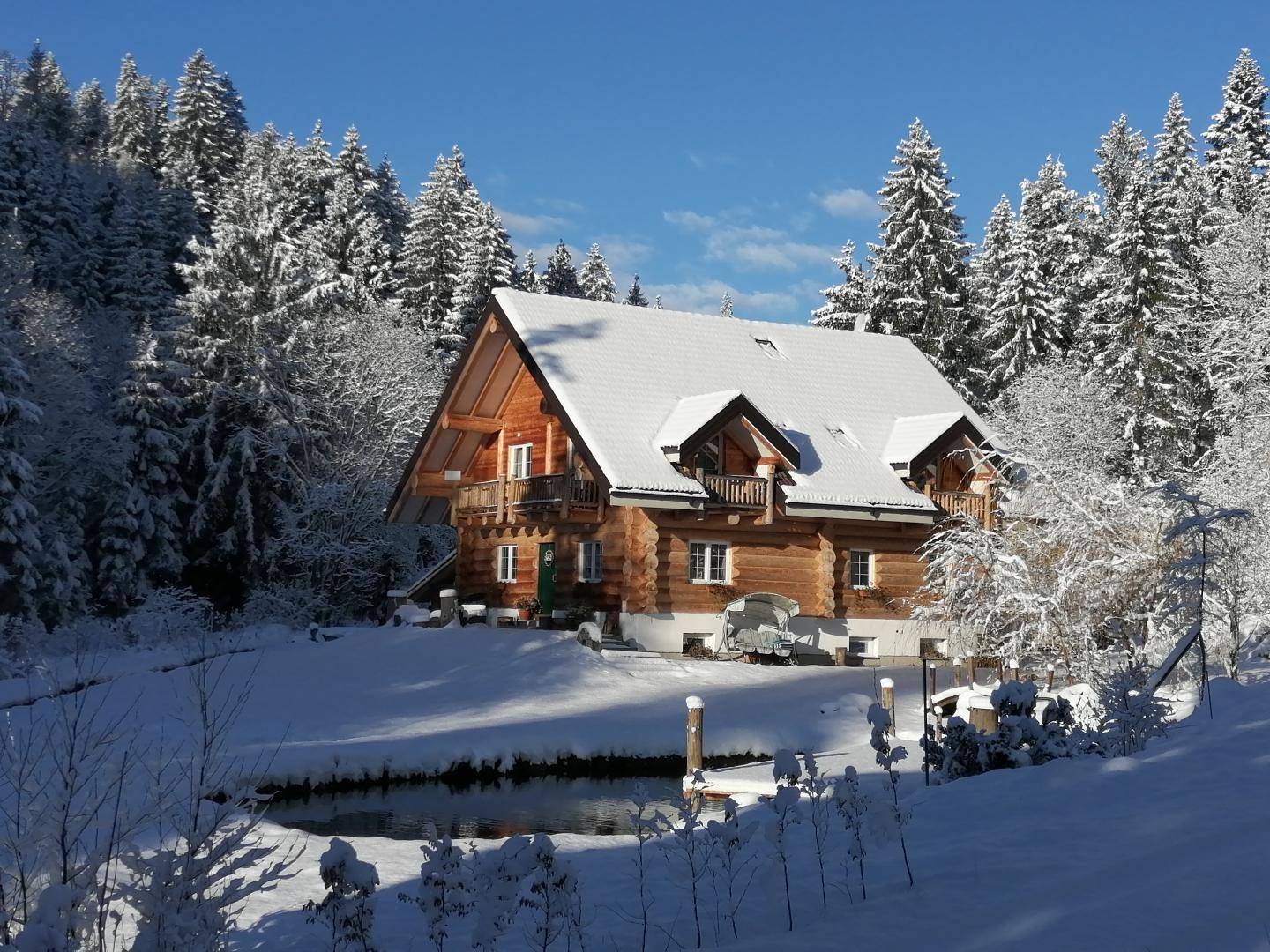Blockhaus im Winter mit verschneitem Garten und teilweise zugefrorenem Teich.